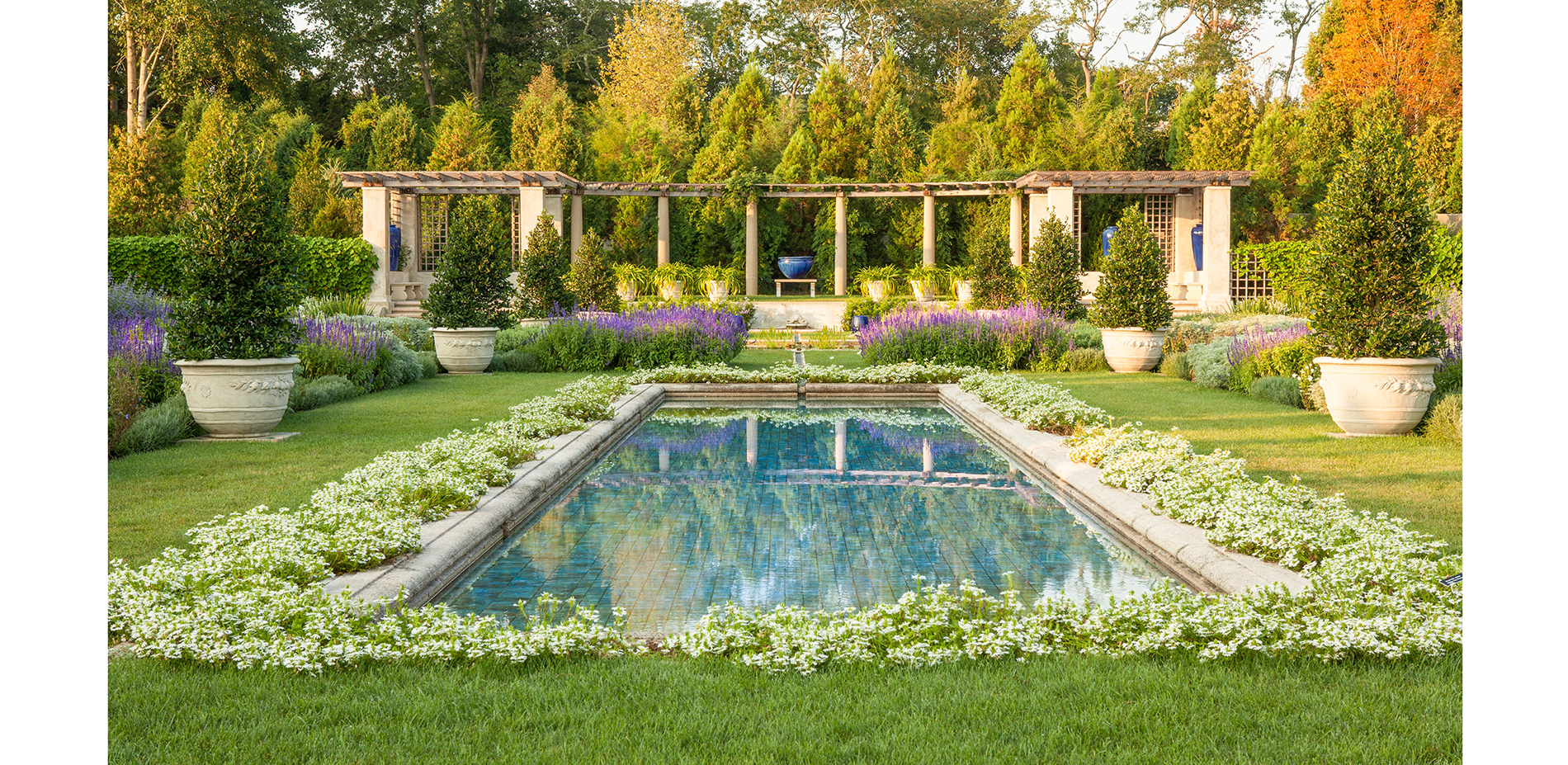 Looking south down the tiled pool towards the pergola, with the space framed by mixed evergreen trees.…