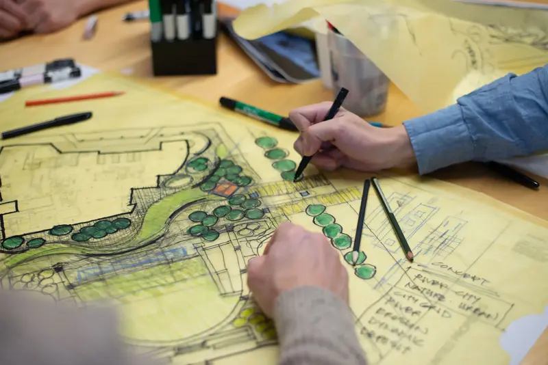 A group of individuals collaborates on an architectural design at a table. The design includes plans of a building, surrounded by green spaces and trees. Pens and rulers are scattered on the table.