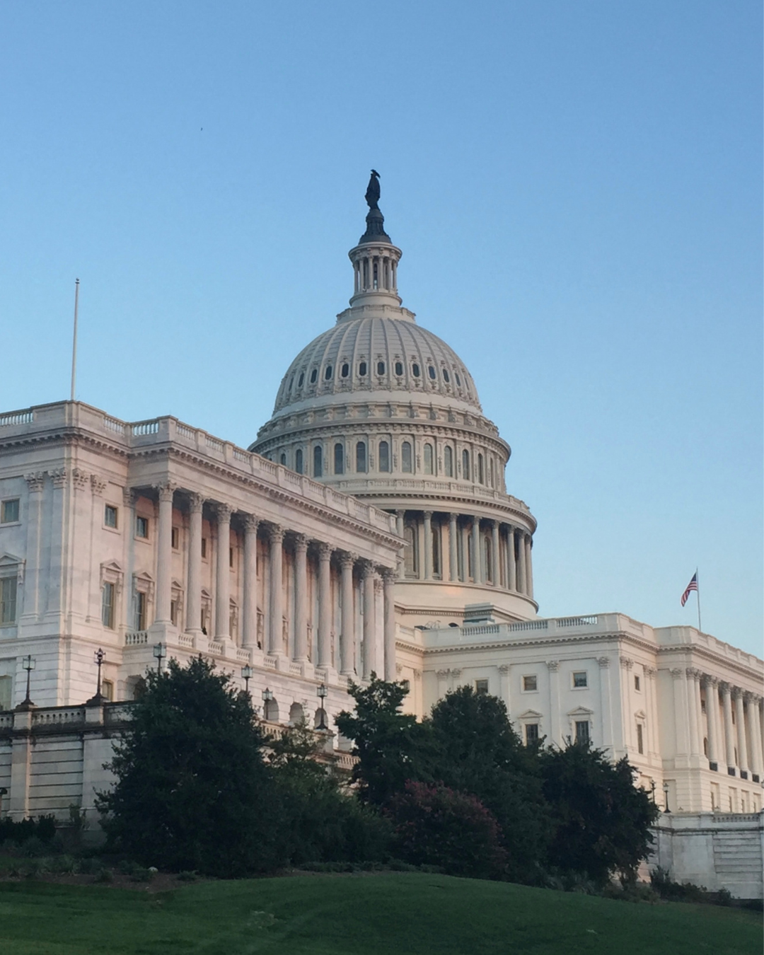 The image shows the United States Capitol building with its iconic dome at dusk. The sky is clear, and the building is surrounded by greenery. An American flag is visible on the right side.