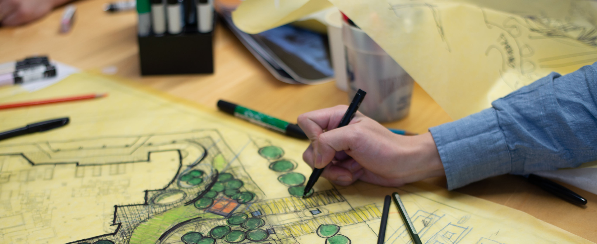 A person is drawing a concept plan on yellow tracing paper with colored pens. The plan includes paths and trees drawn in green. Several pens and drafting tools are scattered on the table.