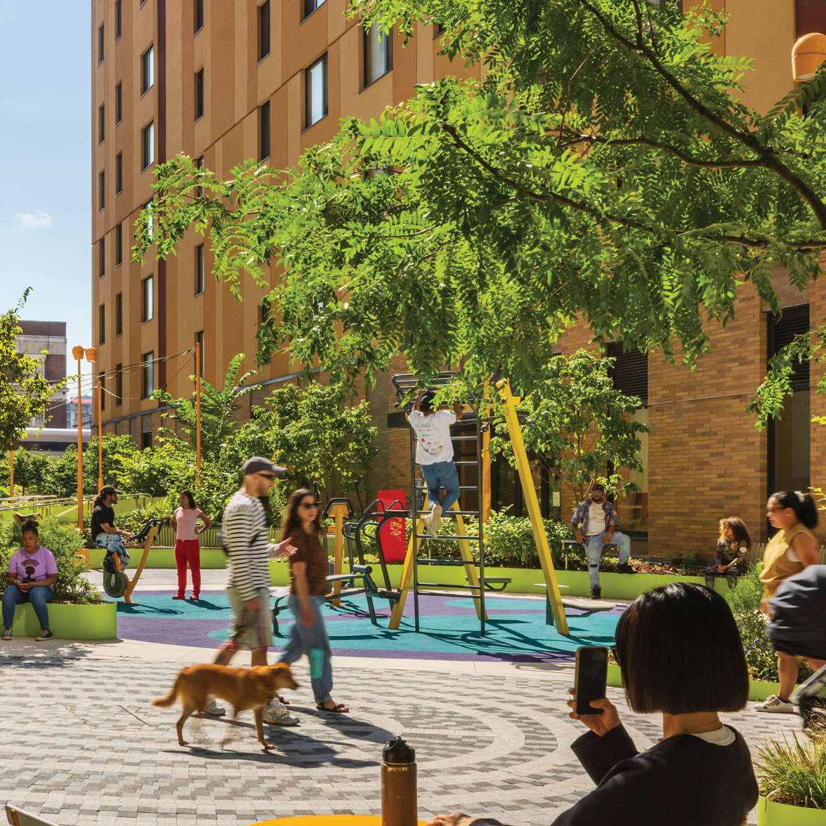A vibrant urban courtyard with people enjoying outdoor activities. The area is surrounded by trees and modern buildings, creating a lively community atmosphere.