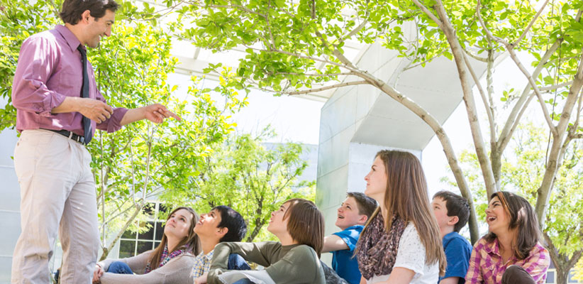 A group of young students sitting on an outdoor deck and watching an instructor who is holding up a leaf